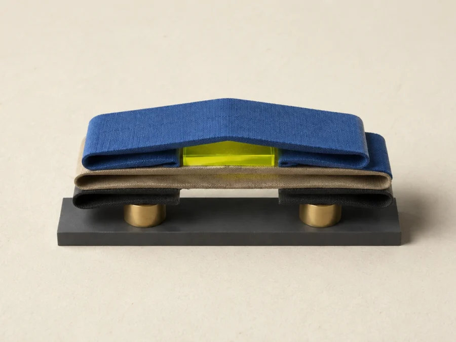 Folded cobalt, sand, and charcoal fabric layers rest on brass supports while a lime acrylic block lifts the top layer.