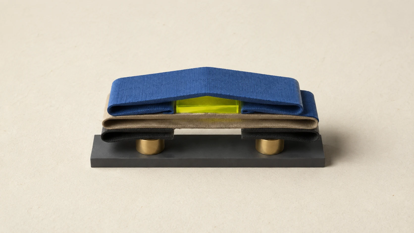 Folded cobalt, sand, and charcoal fabric layers rest on brass supports while a lime acrylic block lifts the top layer.