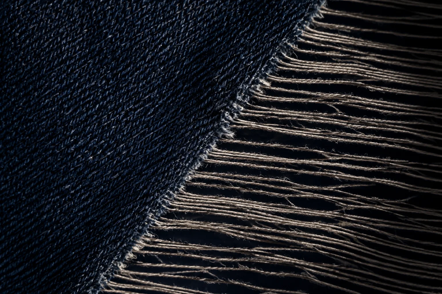 Dark indigo cloth meets loose pale threads along a diagonal edge against a black background.