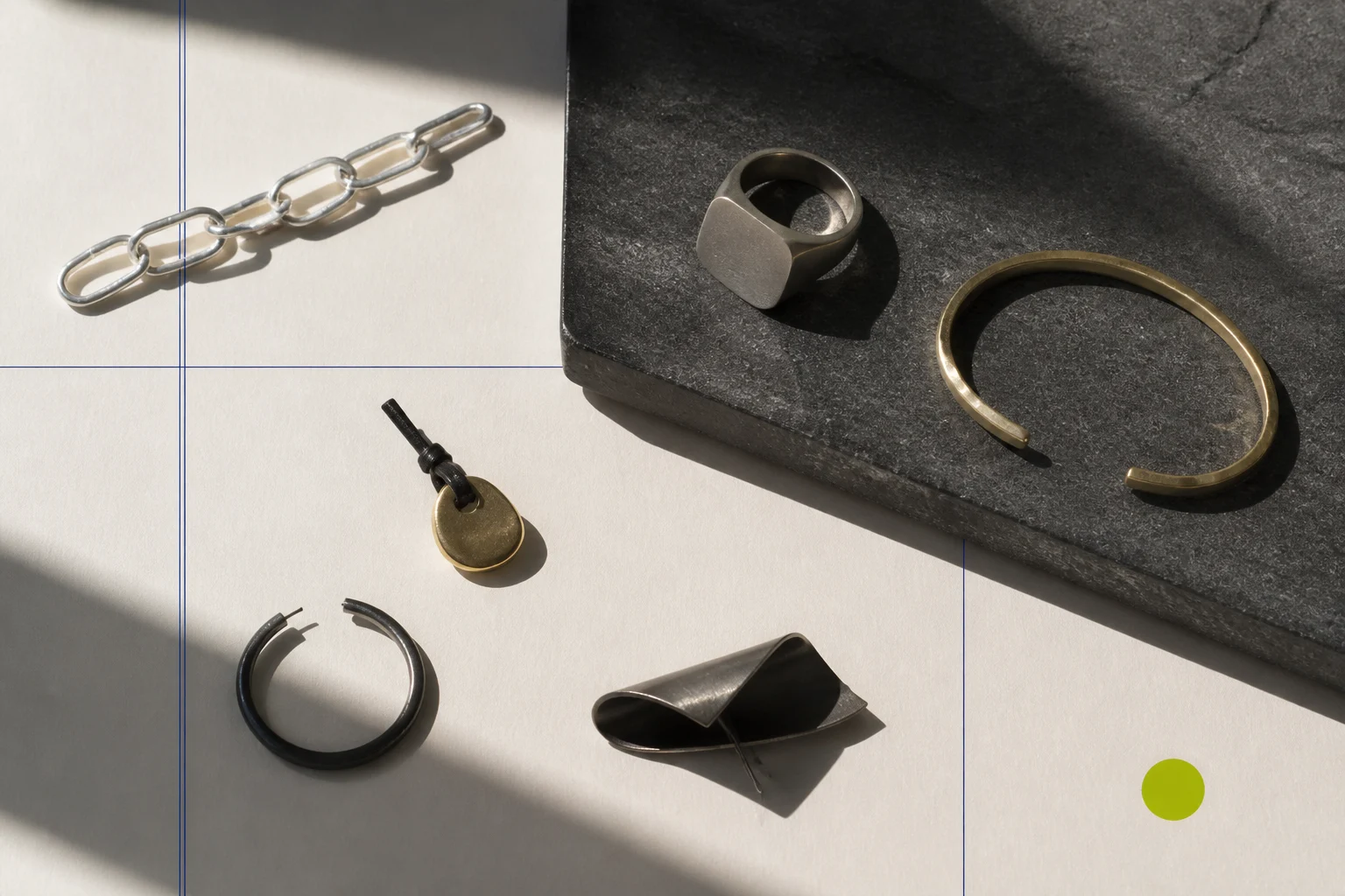 A non-documentary jewelry still life arranges chain, ring, cuff, pendant, earring, and pin forms across graphite, silver, warm paper, cobalt, and one acid-lime marker.